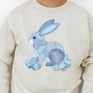 Pode incluir: Camisola cinza claro com um design de coelho patchwork em tons de azul, verde e branco. O coelho tem orelhas grandes e está sentado. A camisola é usada por uma criança.