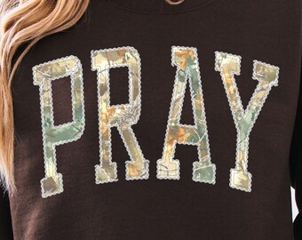 May include: A dark brown sweatshirt with the word "PRAY" in large, textured letters across the front. The letters have a camouflage-like pattern with a white scalloped border. The sweatshirt has long sleeves and a crew neck.