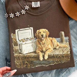 May include: A brown t-shirt featuring a graphic of a golden retriever dog lying on a wooden dock next to a cooler filled with cans. The shirt also has the text "Comfort Colors" on the tag.