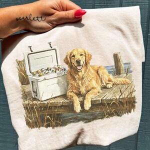 May include: A light beige t-shirt featuring a golden retriever dog lying on a wooden dock next to a white cooler filled with cans. The dog has a happy expression. The scene is set near water with tall grass.