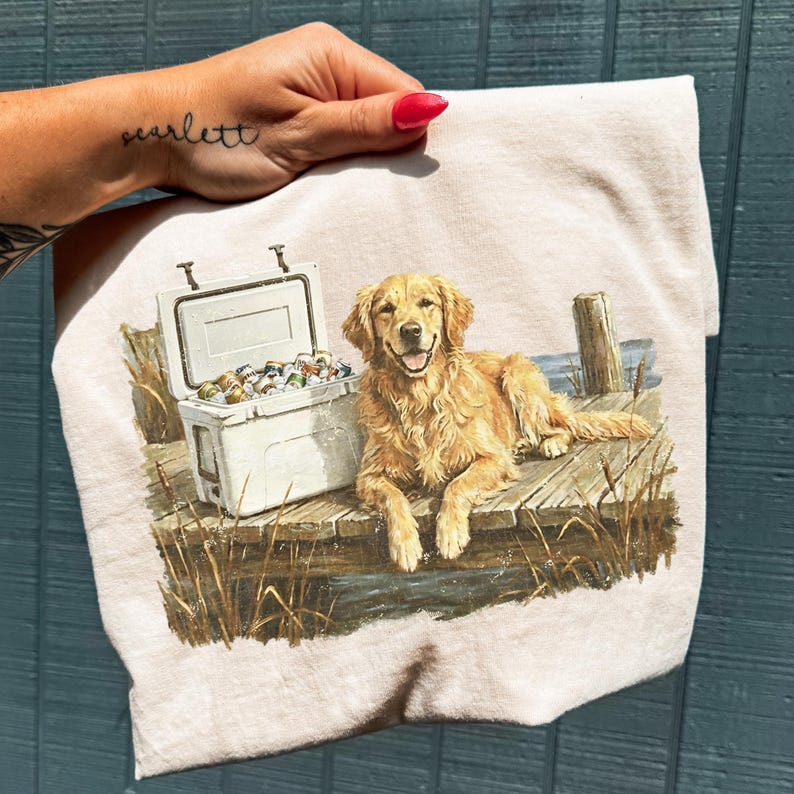 May include: A light beige t-shirt featuring a golden retriever dog lying on a wooden dock next to a white cooler filled with cans. The dog has a happy expression. The scene is set near water with tall grass.