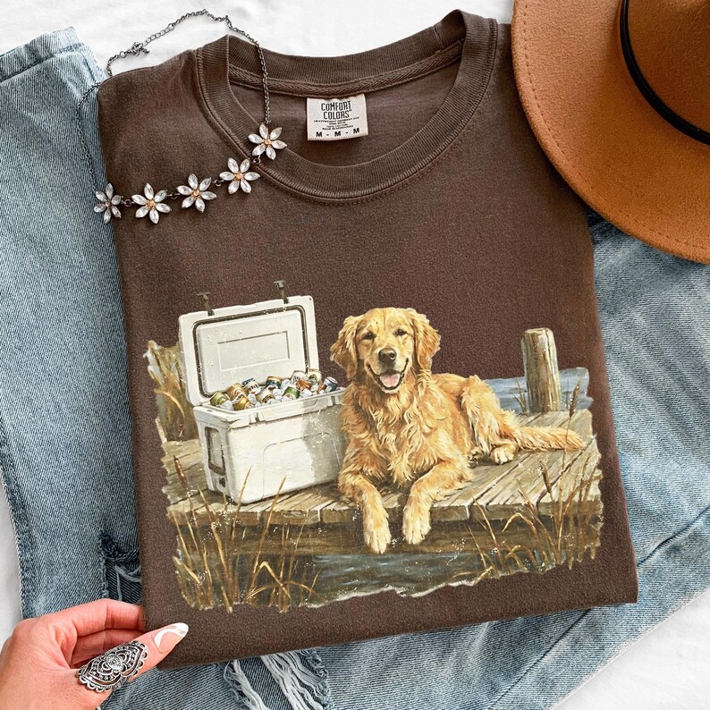 May include: A brown t-shirt featuring a graphic of a golden retriever dog lying on a wooden dock next to a cooler filled with cans. The shirt also has the text "Comfort Colors" on the tag.