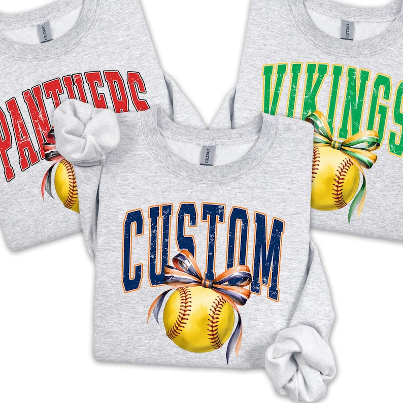 May include: Three light gray sweatshirts with different text and softball graphics. One says "CUSTOM" in navy blue, another says "PANTHERS" in red, and the third says "VIKINGS" in green. Each graphic includes a yellow softball with a bow.