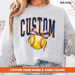 May include: Ash grey sweatshirt with the word "CUSTOM" in navy blue arched lettering. A yellow softball with navy and orange ribbon is centered below the text. The bottom of the sweatshirt has the text "CUSTOM TEAM NAME & TEAM COLORS".
