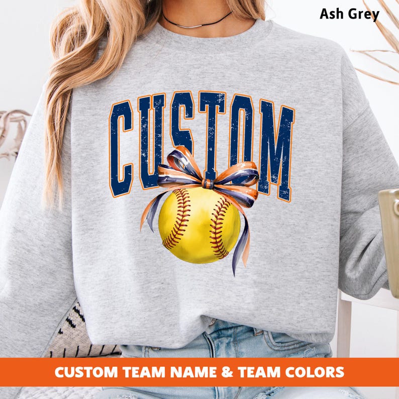May include: Ash grey sweatshirt with the word "CUSTOM" in navy blue arched lettering. A yellow softball with navy and orange ribbon is centered below the text. The bottom of the sweatshirt has the text "CUSTOM TEAM NAME & TEAM COLORS".