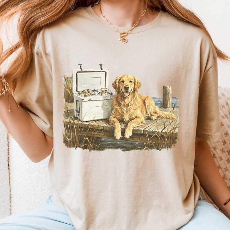 May include: Beige t-shirt featuring a golden retriever dog lying on a wooden dock next to a white cooler filled with colorful items. The dog is smiling. The scene includes water, reeds, and a wooden post.