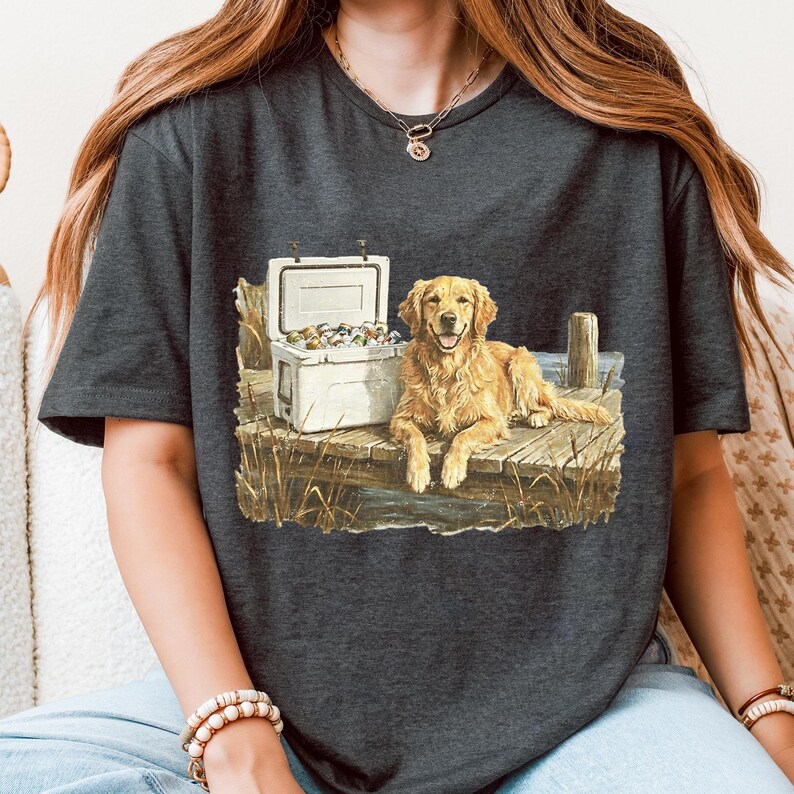 May include: Dark gray t-shirt featuring a golden retriever lying on a wooden dock next to a white cooler. The dog has a happy expression. The graphic includes a detailed illustration of the dog, cooler, and dock.