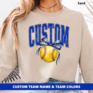 May include: Sand-colored sweatshirt with the word "CUSTOM" in blue, above a yellow softball tied with a blue ribbon. The bottom of the sweatshirt has the text "CUSTOM TEAM NAME & TEAM COLORS" in white on a blue background.