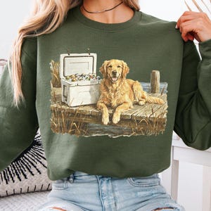 May include: Green sweatshirt featuring a Golden Retriever lying on a wooden dock next to a white cooler filled with items. The dog has a happy expression. The design includes water, reeds, and a wooden post.