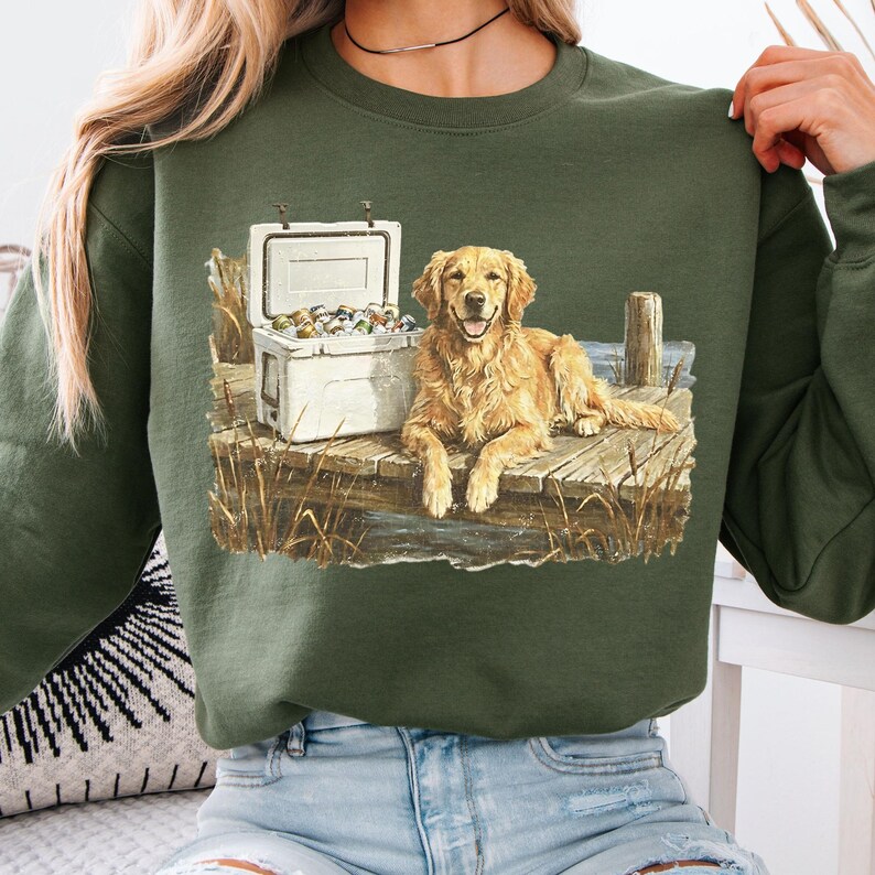 May include: Green sweatshirt featuring a Golden Retriever lying on a wooden dock next to a white cooler filled with items. The dog has a happy expression. The design includes water, reeds, and a wooden post.