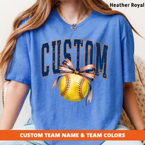 May include: Heather Royal blue t-shirt with the word "CUSTOM" in distressed navy blue lettering. A yellow softball with brown stitching and a navy and orange bow is centered below the word. The text "CUSTOM TEAM NAME & TEAM COLORS" is at the bottom.