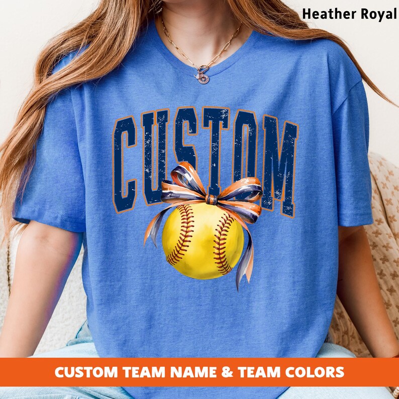 May include: Heather Royal blue t-shirt with the word "CUSTOM" in distressed navy blue lettering. A yellow softball with brown stitching and a navy and orange bow is centered below the word. The text "CUSTOM TEAM NAME & TEAM COLORS" is at the bottom.