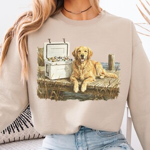 May include: Beige sweatshirt featuring a Golden Retriever dog lying on a wooden dock next to a white cooler. The cooler is open, revealing its contents. The dog is smiling. The design is a watercolor illustration.