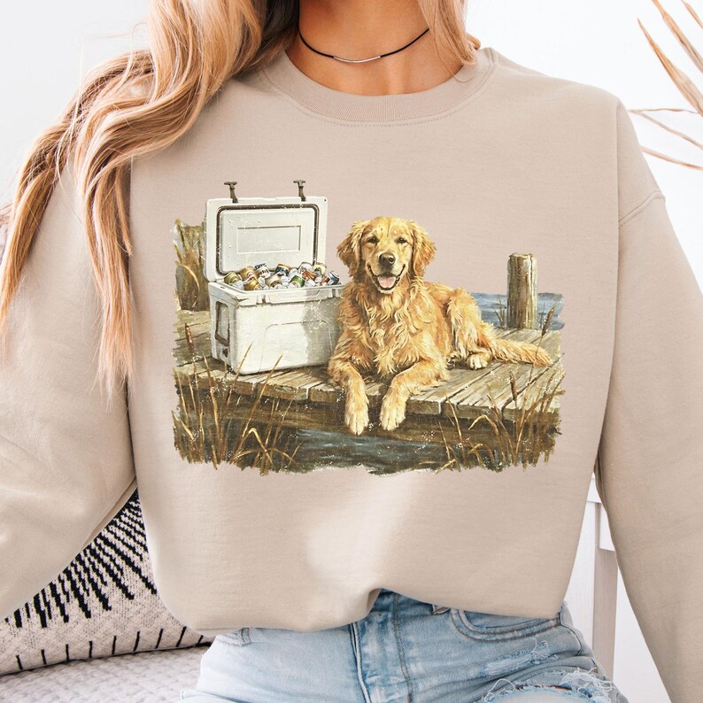 May include: Beige sweatshirt featuring a Golden Retriever dog lying on a wooden dock next to a white cooler. The cooler is open, revealing its contents. The dog is smiling. The design is a watercolor illustration.