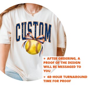 May include: Cream-colored t-shirt featuring the word "CUSTOM" in navy blue, arched lettering. A yellow softball with navy and orange ribbon is centered below the text. The shirt has text that says "AFTER ORDERING, A PROOF OF THE DESIGN WILL BE MESSAGED TO YOU" and "48-HOUR TURNAROUND TIME FOR PROOF."