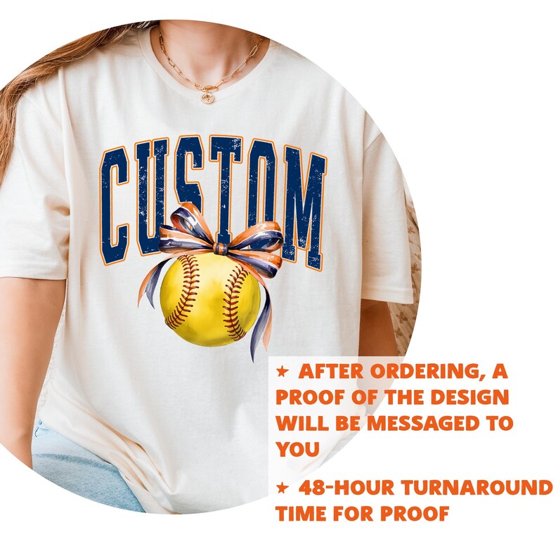 May include: Cream-colored t-shirt featuring the word "CUSTOM" in navy blue, arched lettering. A yellow softball with navy and orange ribbon is centered below the text. The shirt has text that says "AFTER ORDERING, A PROOF OF THE DESIGN WILL BE MESSAGED TO YOU" and "48-HOUR TURNAROUND TIME FOR PROOF."