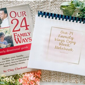 May include: A white and pink devotional notebook with gold lettering that reads "Our 24 Family Ways Copy Work Notebook" and a smaller line of text that reads "Alexa Grammar". The notebook is sitting on a beige woven surface next to a closed book with a red and white cover that reads "Our 24 Family Ways" and "A family devotional guide". The book has a photo of a family on the cover.