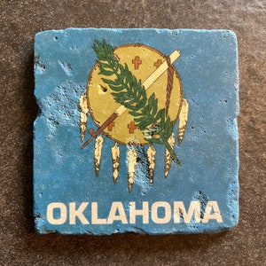May include: Square, blue coaster with the Oklahoma state seal. The seal features a central shield with a star, olive branch, and Native American symbols. The word "OKLAHOMA" is printed in white at the bottom.
