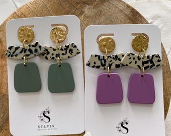 May include: Two pairs of handmade earrings on white cards. Each earring features a gold-colored disc, a cream-colored section with black speckles, and a square-shaped bottom in either green or purple. The cards display the brand name "SYLVIS".