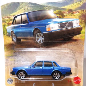 Op de afbeelding: Een blauwe gegoten Volvo sedan speelgoedauto. De speelgoedauto staat op een kartonnen achtergrond met een landschappelijke achtergrond. De auto heeft zwarte wielen en een gedetailleerde grille. De verpakking bevat de tekst "METAL LIFETIME" en "3+ MATTEL".