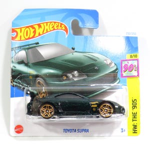 Puede incluir: Un coche de juguete Hot Wheels Toyota Supra verde con llantas doradas, empaquetado en una tarjeta azul y blanca. El embalaje presenta el logotipo de Hot Wheels y el texto "HW: The '90s". El coche está etiquetado como "Toyota Supra".