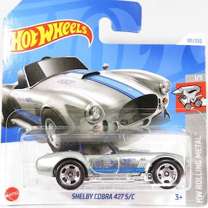Puede incluir: Un coche de juguete de metal fundido a presión Hot Wheels de color plateado que representa un Shelby Cobra 427 S/C. El coche tiene rayas de carreras azules y un interior negro. El coche está sobre un fondo azul y blanco con el logotipo de Hot Wheels y las palabras "HW Rolling Metal".