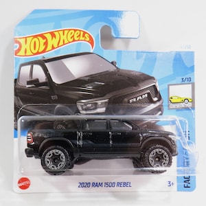 Könnte beinhalten: Ein schwarzes Spielzeugauto vom Typ Pick-up Ram 1500 Rebel 2020 in einer blauen und weißen Hot Wheels-Verpackung. Das Auto hat schwarze Räder mit silbernen Akzenten und die Worte "RAM" und "REBEL" sind an der Seite des LKWs sichtbar.