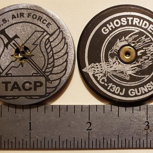 May include: Two round metal coins. The first coin is silver with a black and white image of a winged sword and the text "U.S. AIR FORCE" and "TACP". The second coin is black with a silver image of a skull with flames and the text "GHOST RIDER AC-130J GUNSHIP".