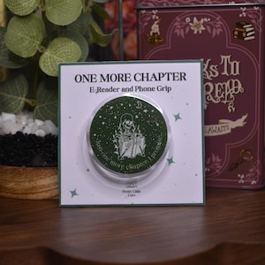 Peut inclure: Un support de téléphone vert et blanc avec le texte "ONE MORE CHAPTER" et "E-Reader and Phone Grip". Le support présente un squelette lisant un livre sous un croissant de lune et des étoiles, avec le texte "just one more chapter, I promise".