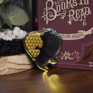 Könnte beinhalten: Ein herzförmiger Emaille-Pin mit einem schwarzen und goldenen Drachenschuppen-Design. Der Pin befindet sich vor einem verschwommenen Hintergrund mit einer dekorativen Box mit den Worten "Books to Read" und einem Terrarium.