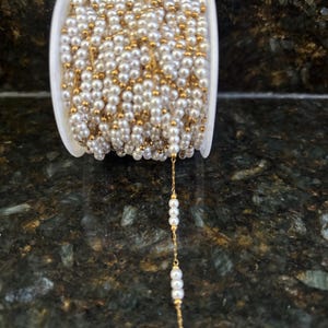 Puede incluir: Un carrete de cadena delicada con pequeñas cuentas esféricas blancas intercaladas con diminutas cuentas de metal en tono dorado. La cadena está enrollada firmemente alrededor de un carrete cilíndrico de plástico blanco, con un hilo que cuelga.