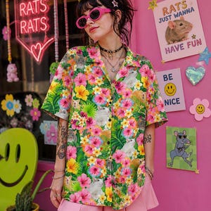 Könnte beinhalten: Ein kurzärmeliges Hemd mit einem floralen und Ratten-Print. Das Hemd zeigt rosa, gelbe und grüne Blumen, mit weißen und braunen Ratten. Der Hintergrund umfasst rosa Wände, Neon-Schilder mit "RATS" und Poster mit Ratten-Grafiken.