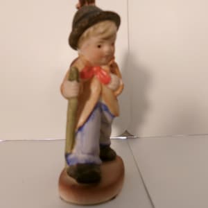 May include: A ceramic figurine of a young boy in a tan coat, blue pants, and a brown hat. He holds a green staff and has a red bow tie. The figurine is standing on a brown oval base.