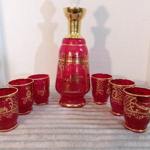 May include: A ruby red glass decanter set with six matching glasses. The decanter has a gold-colored stopper and gold trim. The glasses and decanter are decorated with gold filigree and architectural designs.
