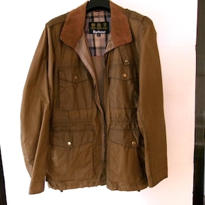May include: A brown waxed cotton jacket with a corduroy collar and a plaid lining. The jacket has multiple pockets, a zip and snap closure, and the Barbour logo on the collar. The jacket is hanging on a black hanger.