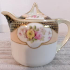 May include: A white porcelain creamer with a lid, featuring a floral design with pink and yellow roses. The creamer has a peach-colored band with gold trim and a black and gold patterned top.