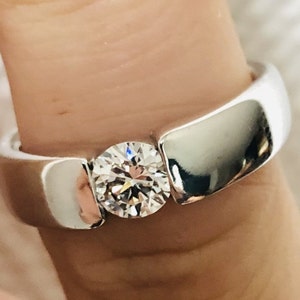 Op de afbeelding: Een zilveren ring met een grote, ronde, heldere diamant in het midden. De band is dik en heeft een gladde, gepolijste afwerking.