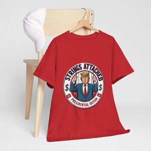 Puede incluir: Camiseta roja con un gráfico circular blanco que presenta una imagen de dibujos animados de Donald Trump con el texto "Strings Attached Presidential Edition".