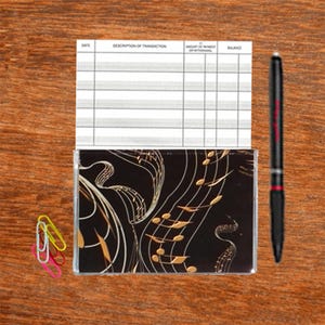 May include: A checkbook with a black cover featuring gold musical notes and staff lines. A black pen with red accents and three colorful paper clips are also on the wooden surface.
