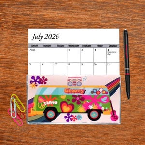 May include: A July 2026 calendar with a groovy, psychedelic van design. The van is green, pink, and orange with peace signs, flowers, and hearts. The calendar is on a wooden table with a pen and paper clips.