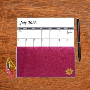 May include: A July 2026 calendar with a pink glitter cover and a small yellow flower sticker. The calendar is on a wooden surface with a pen and paper clips.