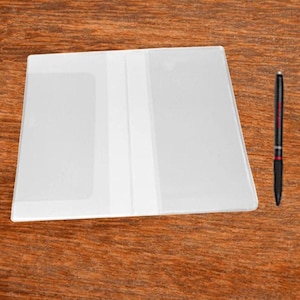 May include: A clear, plastic document holder with a white spine, lying on a wooden surface. A black pen with red accents rests beside the holder. The holder is rectangular and appears to be empty.