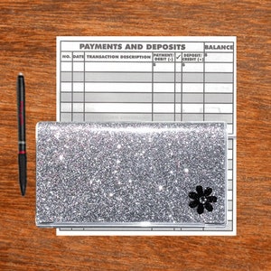 May include: A silver glitter checkbook cover with a black flower design. The cover is on top of a white checkbook with the words "Payments and Deposits" and "Balance" printed on it.