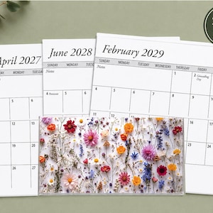 May include: A set of three white paper calendar pages for April 2027, June 2028, and February 2029, featuring black text and grid lines. A rectangular clear acrylic block with a colorful pressed wildflower pattern sits in front of the pages.