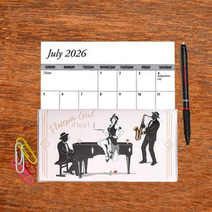 May include: A calendar for July 2026 with a decorative image of a flapper girl, a pianist, and a saxophone player. The calendar is on a wooden surface, with a black pen and colorful paper clips.