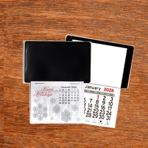 May include: A black desk calendar with a white calendar insert, displaying the months of December 2025 and January 2026. The December calendar has snowflake designs and the words "Happy Holidays". The calendar is on a wooden surface.