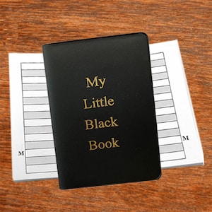 May include: A small, black notebook with the gold text "My Little Black Book" on the cover. The notebook is open, revealing lined pages. The background is a wooden surface.
