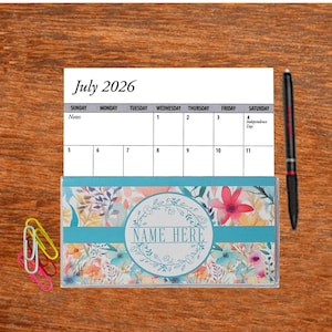 May include: A July 2026 calendar with a floral design checkbook cover. The checkbook cover has a blue background with a wreath of flowers and the text "NAME HERE".