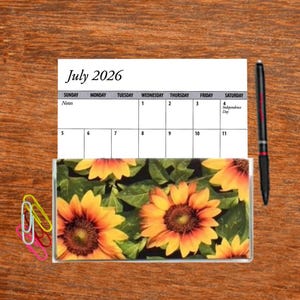 May include: A July 2026 calendar with a sunflower design. The calendar is printed on white paper and has a black and white grid. The sunflower design is a close-up of a field of sunflowers.