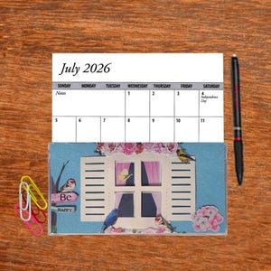 May include: A July 2026 calendar with a decorative window design, featuring birds and floral accents. The calendar is accompanied by a black pen and colorful paper clips. The words "Be Happy" are visible.